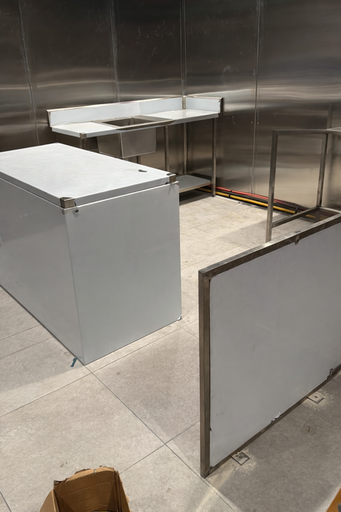 “Indoor stainless steel workspace with reflective metal walls, tiled floor, and three stainless steel worktables; cables neatly arranged along the wall and a cardboard box with tools on the floor.”
It’s concise, descriptive, and includes relevant keywords for your industry. Would you like me to tailor it for Google image SEO (for example, adding ‘Sydney stainless steel fabrication workspace’)?