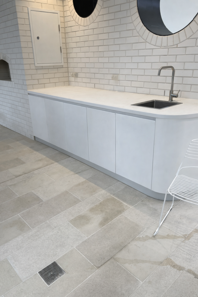 “Modern white benchtop with stainless steel sink and tiled backsplash in a clean indoor space, featuring a wire-frame chair and beige tiled floor.”