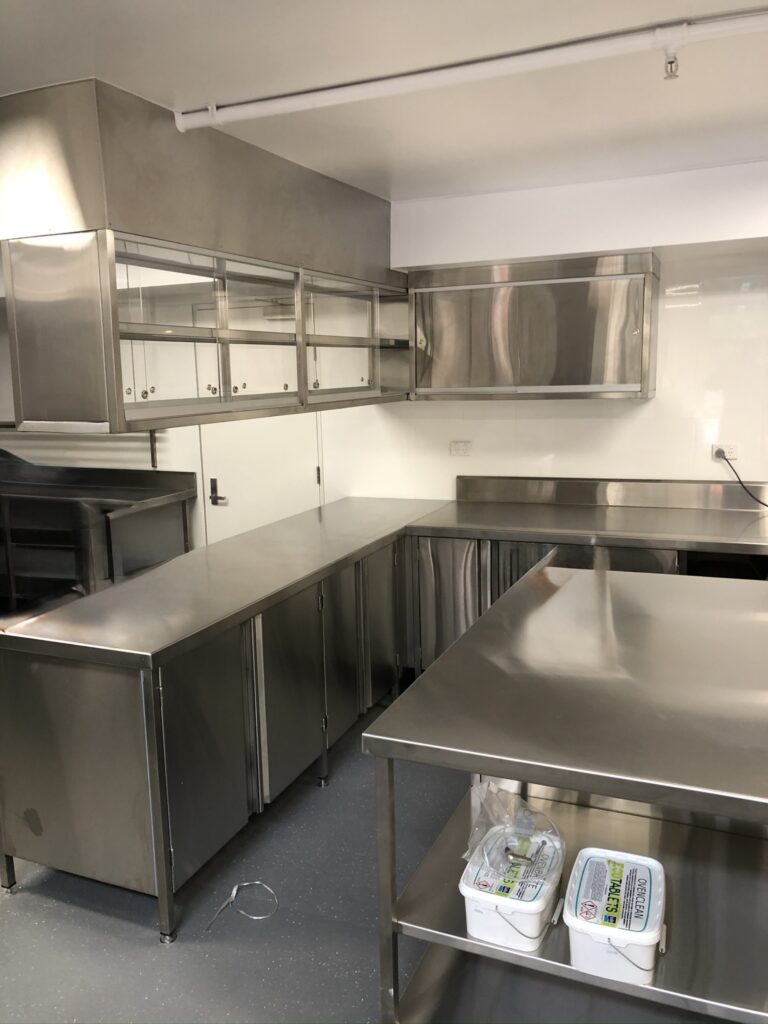 Commercial kitchen fitted with custom 304 stainless steel benches, cabinets, and overhead storage, showing clean durable food preparation surfaces